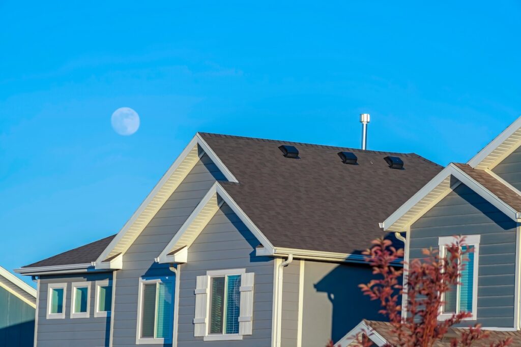 Sunlit home with dark pitched roof over horizontal gray wall siding and windows. Exterior view of house at a neighborhood with blue sky background on a sunny day.
