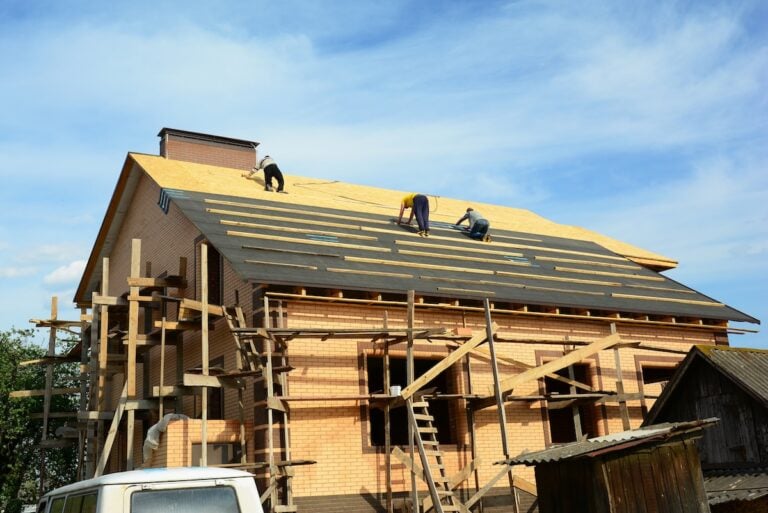 Roofing contractors are installing underlayment, water-resistant, waterproof protection barrier on the roof deck of a large brick house construction with scaffoldings before asphalt shingles covering.