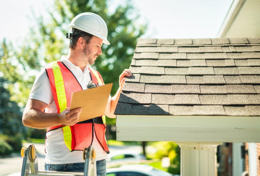 roof inspection after storm A man with hard hat standing on steps inspecting house roof