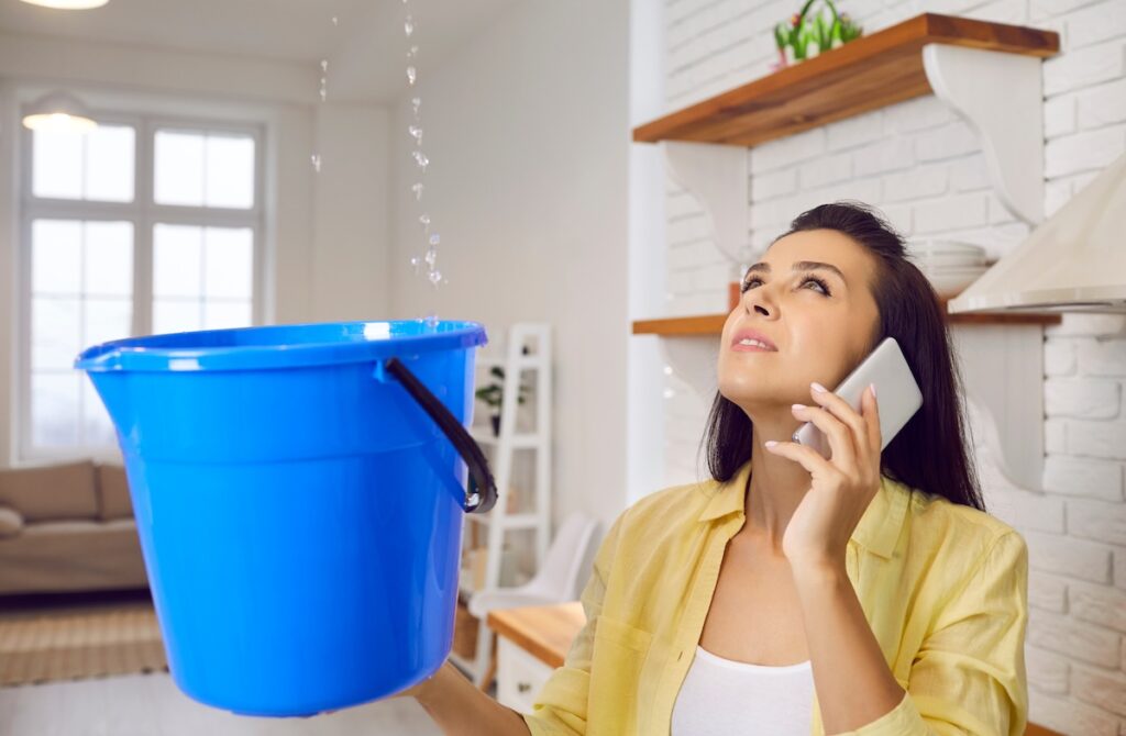 Sad unhappy frustrated young lady speaking to repair service while holding bucket and looking up at water falling down from damaged ceiling in newly renovated kitchen interior. House roof leak concept